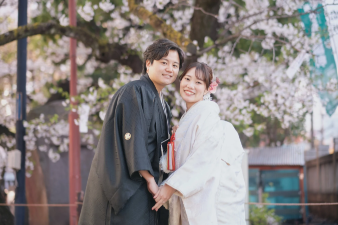 満開の桜の下、和装姿の夫婦が手を取り合い、カメラに向かって笑顔を見せています。