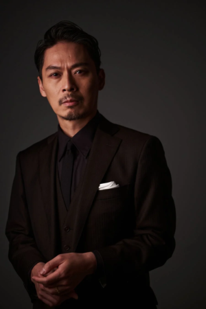 暗い背景を背に、黒いスーツとネクタイを着用した男性のポートレート。口ひげと顎ひげを蓄え、真剣な表情でカメラを見つめている。