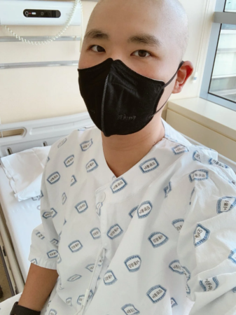 坊主頭の人物が黒いマスクと病院着を着用し、病院のベッドに座っている様子。病院着には「서울대학교병원」（ソウル大学病院）のロゴが見える。入院中の患者の姿を捉えた写真です。