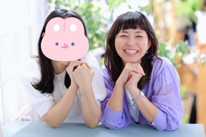 テーブルを挟んで座る2人の女性が写っています。一人は満面の笑みを浮かべており、もう一人の顔はピンクのウサギのマスクで隠されています。和やかな雰囲気の一枚です。