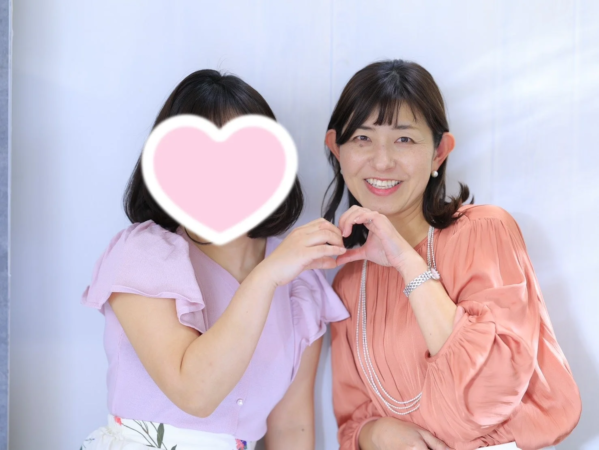 二人の女性が並んでおり、一人は笑顔で両手でハートのポーズを作っています。もう一人の女性の顔はハートのスタンプで隠されており、仲睦まじい雰囲気が伝わります。