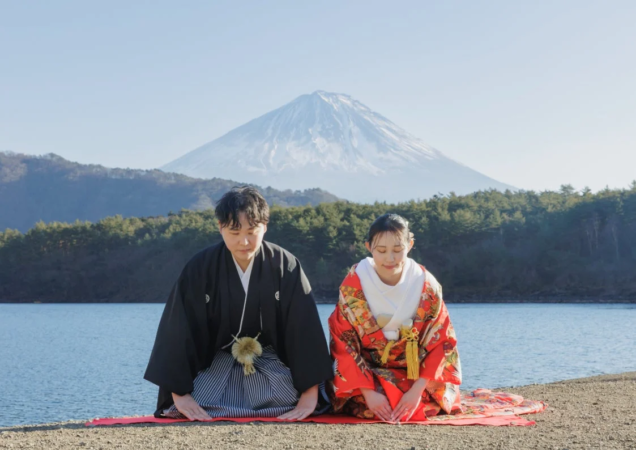 富士山と湖を背景に、和装のカップルが跪いてお辞儀をしている写真です。男性は黒紋付袴、女性は赤の色打掛を着用しており、結婚式の前撮りか記念撮影と思われます。日本の美しい自然と伝統的な衣装が調和した一枚です。