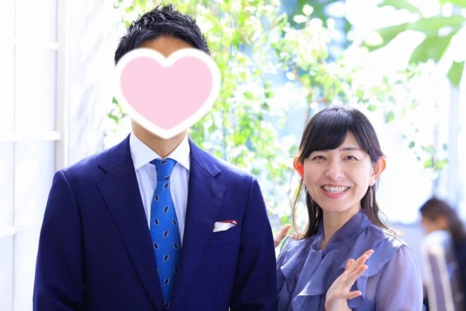 青いスーツ姿の男性（顔はハートで隠されている）と、笑顔でカメラを見つめる紫色のブラウスを着た女性のツーショットです。背景には緑が写り込んでおり、明るく和やかな雰囲気が感じられます。