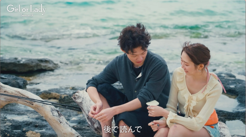 海辺の岩場に座る男女。女性が男性に何かを手渡し、二人は笑顔でリラックスした雰囲気。「Girl or Lady Season 3」の一場面で、釣り竿も見え、穏やかな海辺でのデートシーンを表している。