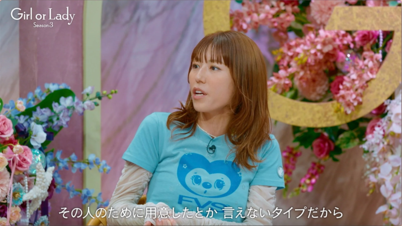 「Girl or Lady Season 3」という番組に出演している女性が、花で飾られたスタジオで話している様子です。青いTシャツを着ており、画面下には「その人のために用意したとか 言えないタイプだから」という日本語の字幕が表示されています。