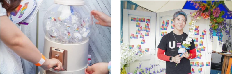 虹色の装飾が施されたイベント会場で、笑顔の男性が花束を持ち、人々がガチャガチャを楽しんでいます。多様性を祝うプライドイベントの様子が伺えます。