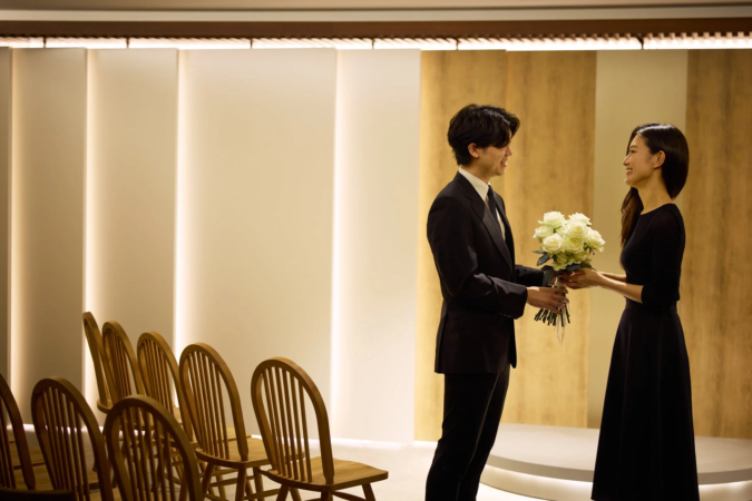 スーツを着た男性が黒いドレスの女性に白いバラの花束を渡しており、二人とも笑顔で向き合っています。木製の椅子が並べられたエレガントな室内で、プロポーズや結婚式、記念日などの特別な瞬間を捉えたようです。