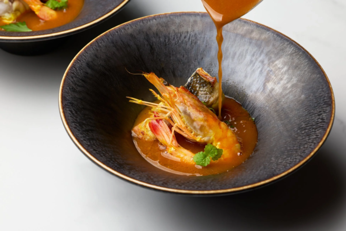 黒い器に盛られた海老と魚料理に、温かいオレンジ色のソースが注がれている瞬間を捉えた写真です。ミントの葉が添えられ、洗練された一皿が表現されています。