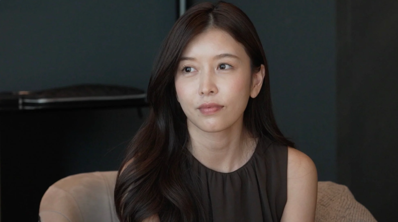ロングヘアの日本人女性が、落ち着いた表情でやや斜め上を見つめているポートレート写真です。ダークカラーのノースリーブの服を着用し、背景は暗くシンプルな室内です。