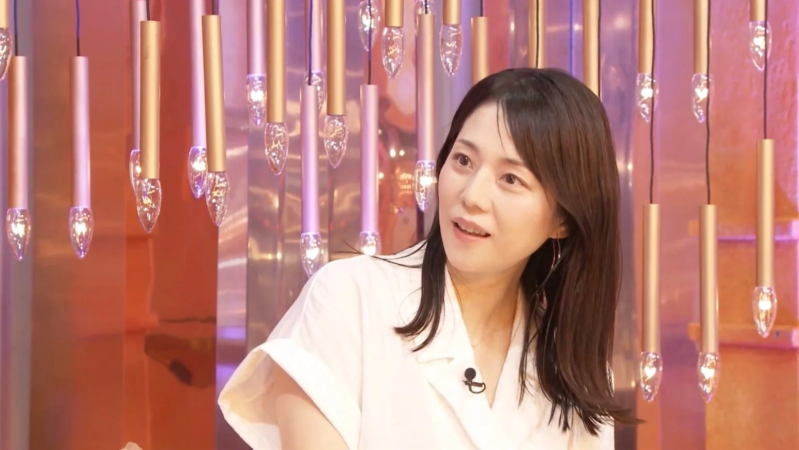 白いシャツを着た女性が、背景に多数の吊り下げ式照明があるスタジオのような場所で、マイクを付けて穏やかな表情で話している様子を捉えた画像です。